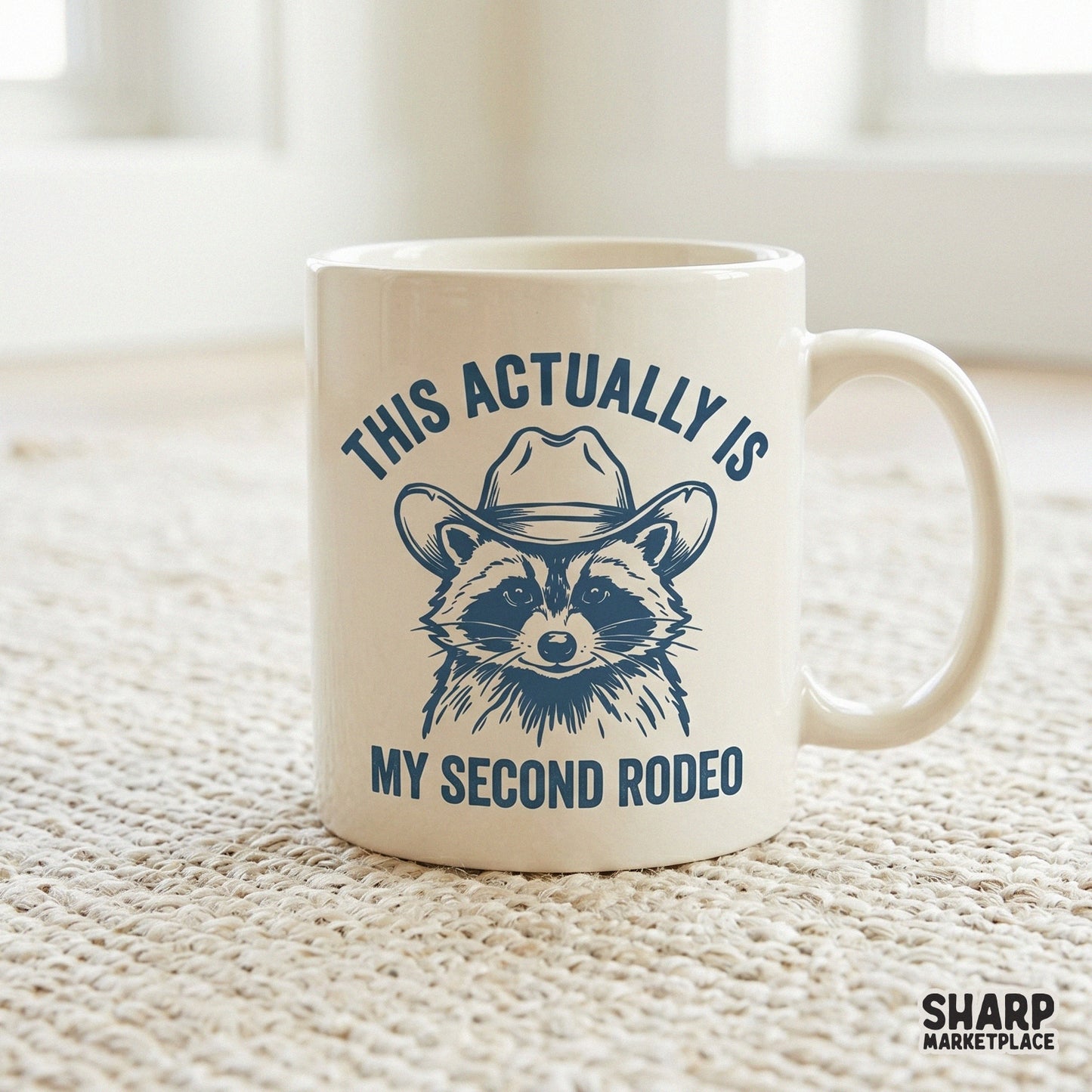 A white coffee mug featuring a slate blue graphic of a raccoon in a cowboy hat with the funny quote This Actually Is My Second Rodeo for western decor.