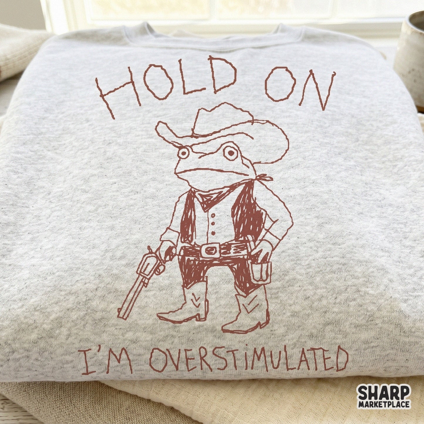 A light gray heather sweatshirt featuring a reddish-brown line art design of a frog sheriff wearing a cowboy hat, vest, and boots with the quote Hold On I'm