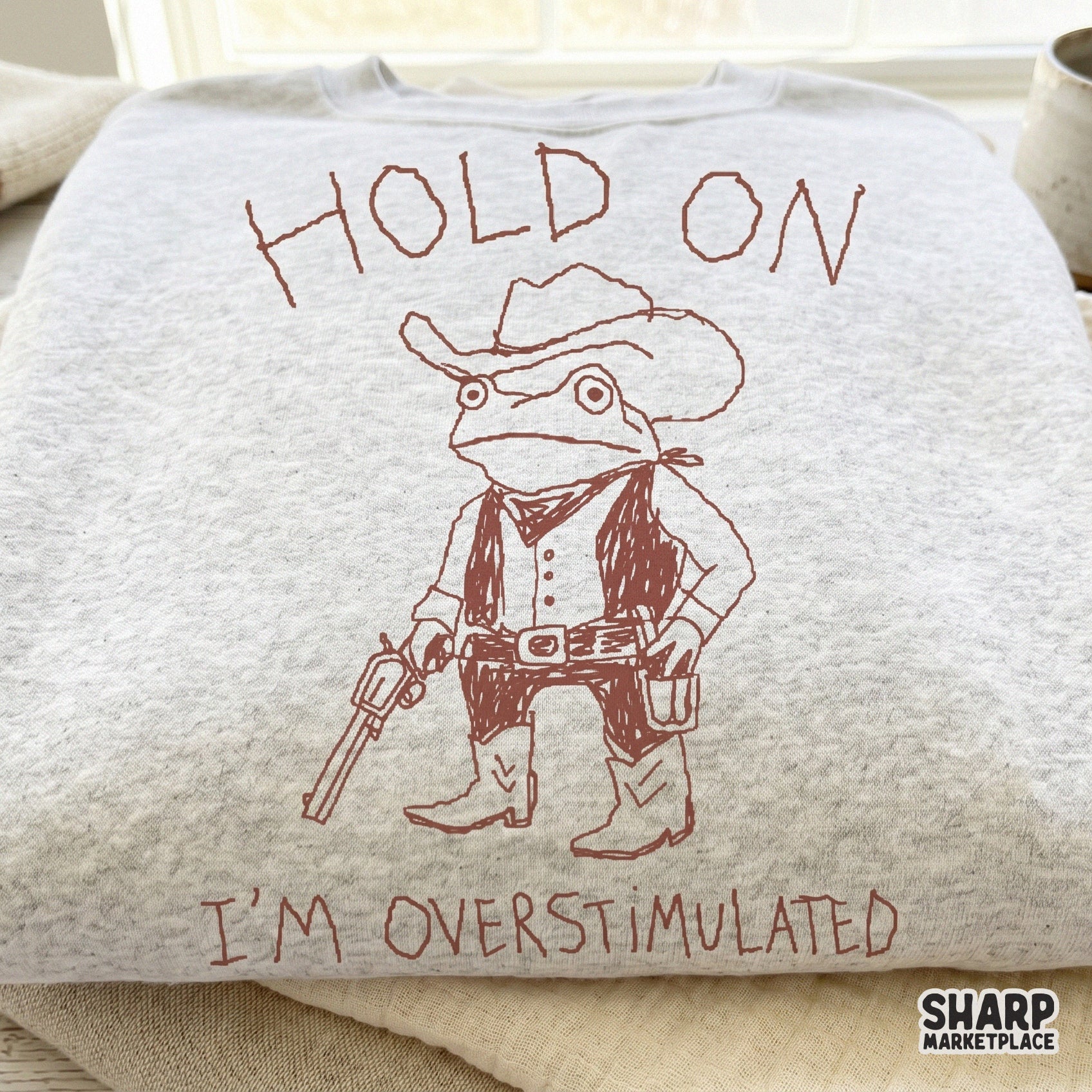 A light gray heather sweatshirt featuring a reddish-brown line art design of a frog sheriff wearing a cowboy hat, vest, and boots with the quote Hold On I'm