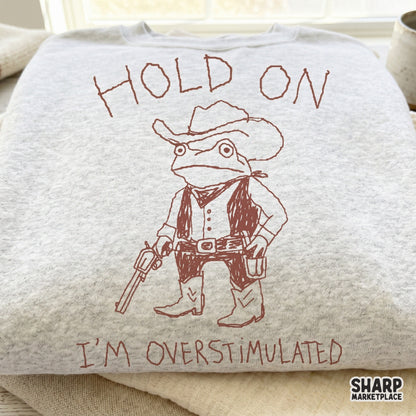A light gray heather sweatshirt featuring a reddish-brown line art design of a frog sheriff wearing a cowboy hat, vest, and boots with the quote Hold On I'm