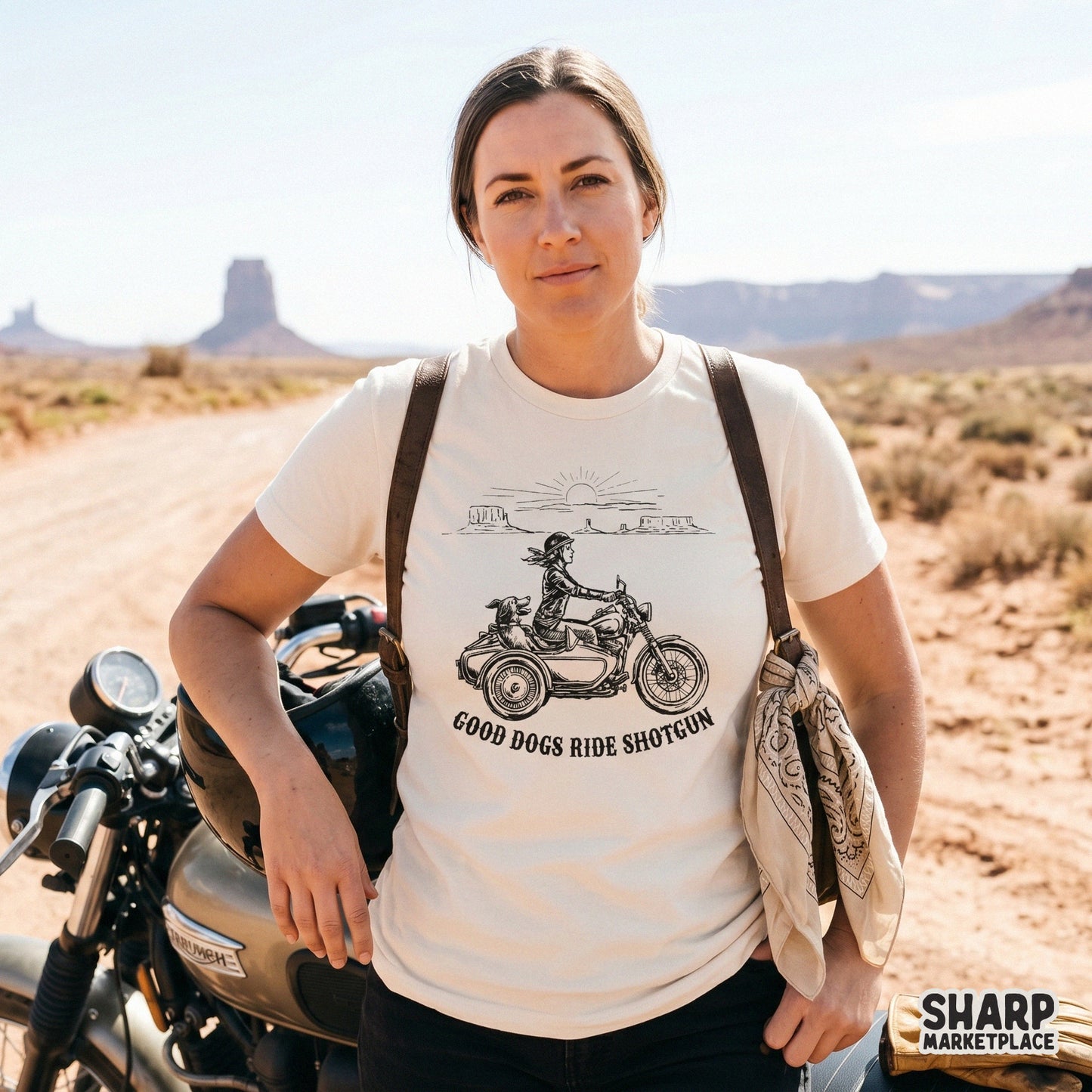 A woman wearing a cream t-shirt featuring a retro black line art design of a biker and her dog in a sidecar, set against a desert mesa horizon.