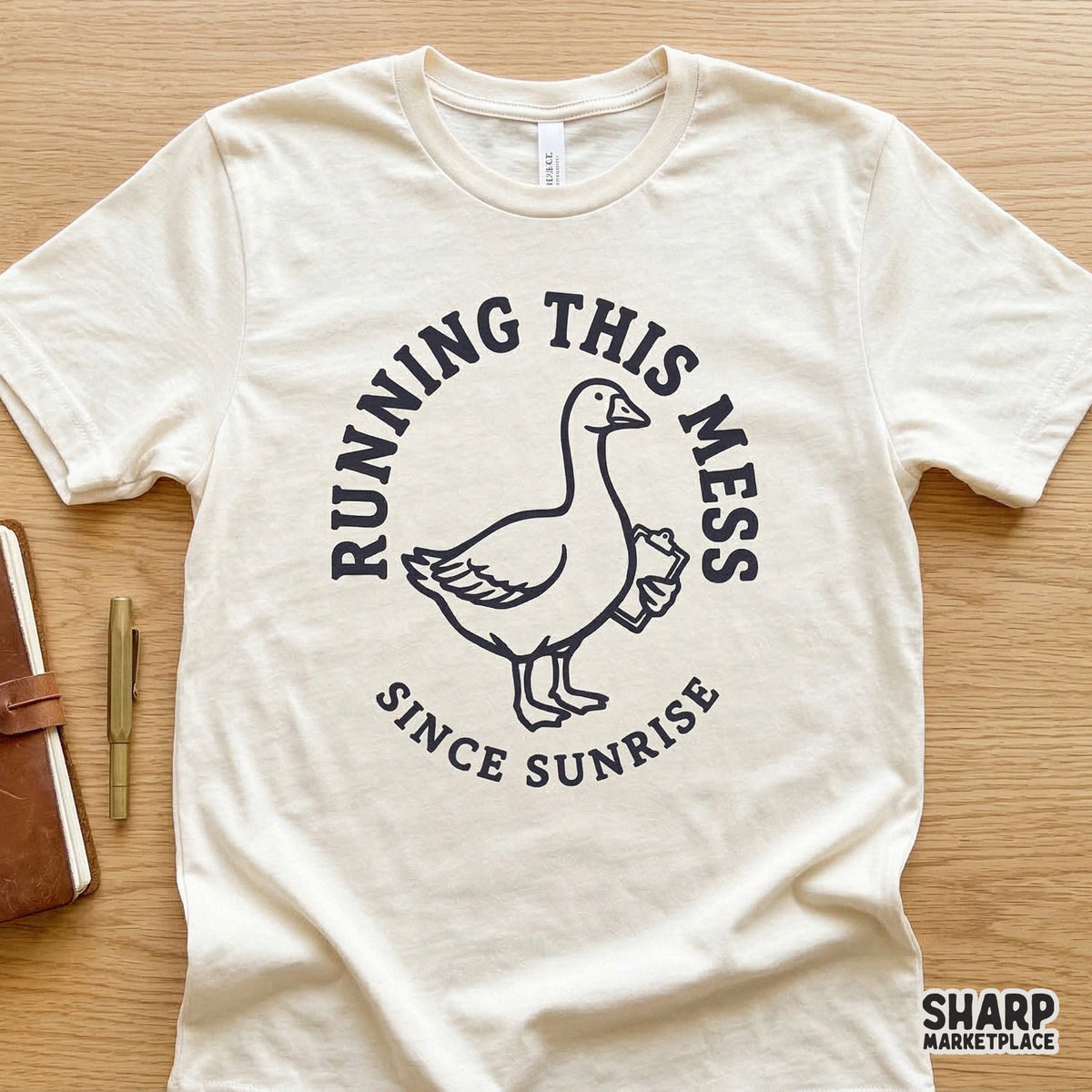 A cream-colored t-shirt featuring a navy blue graphic of a goose with a clipboard and the quote Running This Mess Since Sunrise in a circular layout.