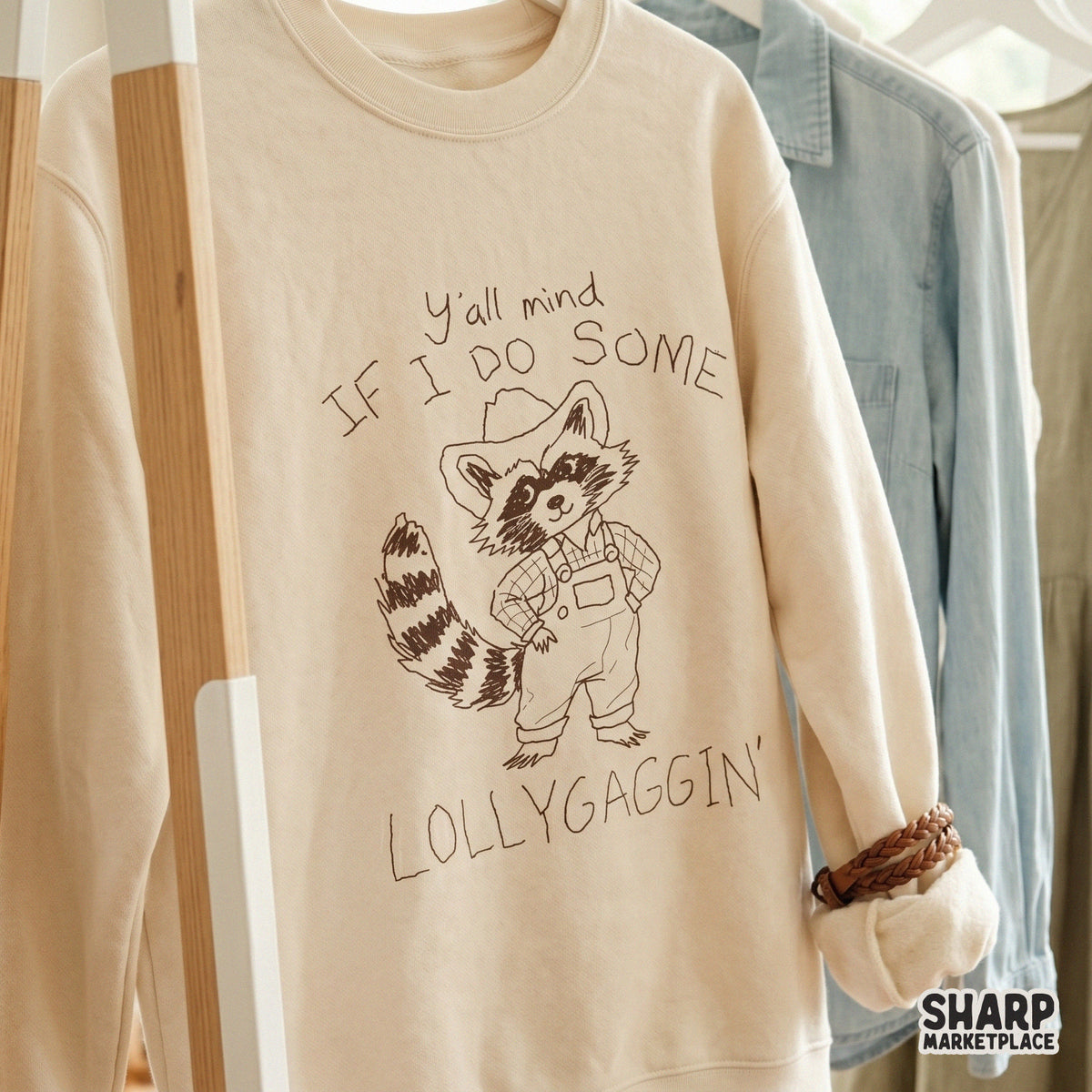 Cream colored sweatshirt featuring a rustic sketch of a raccoon in western overalls and a cowboy hat with the funny quote Y'all Mind If I Do Some Lollygaggin'.