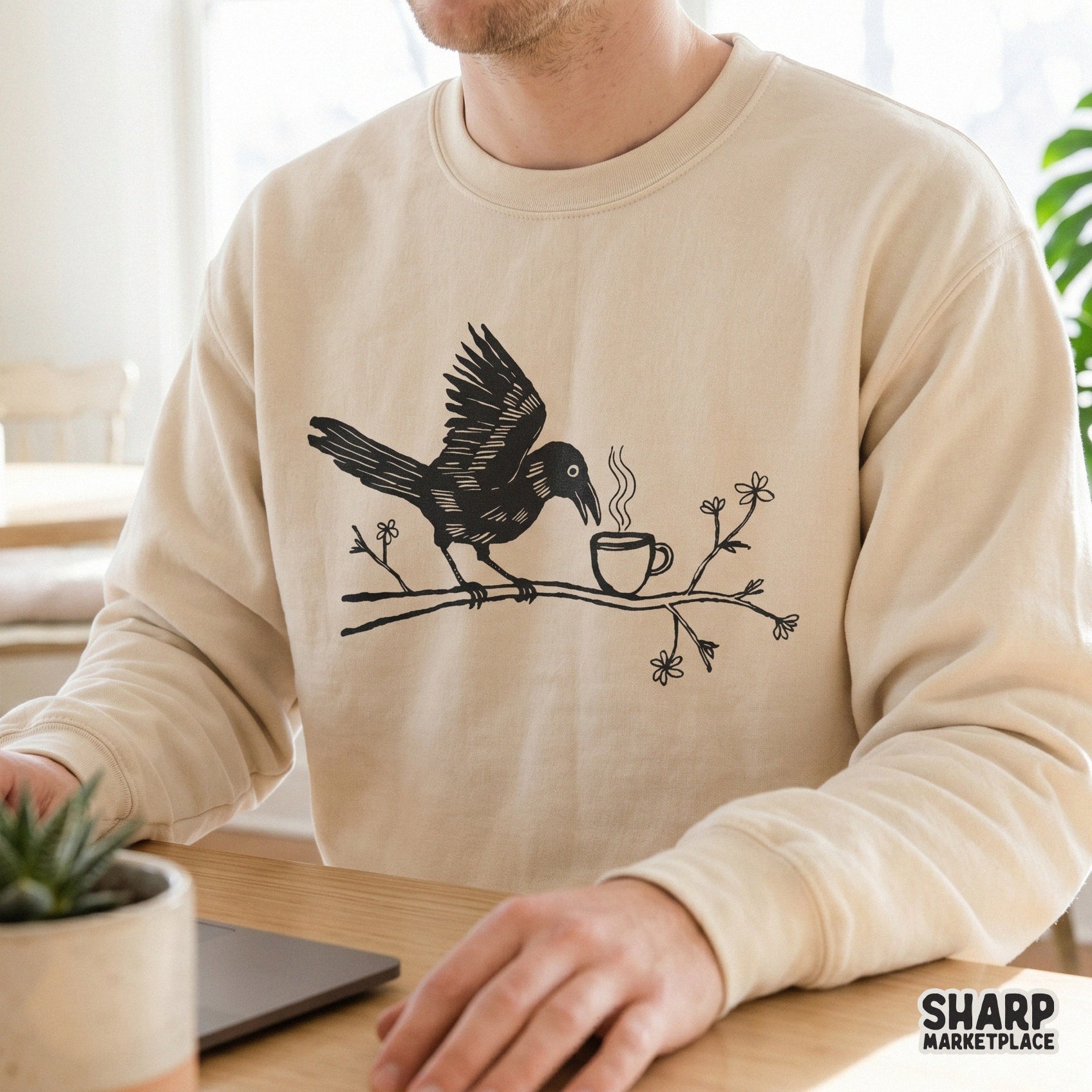 A beige sweatshirt featuring a black hand-drawn design of a crow perched on a branch with flowers, reaching toward a steaming cup of coffee.