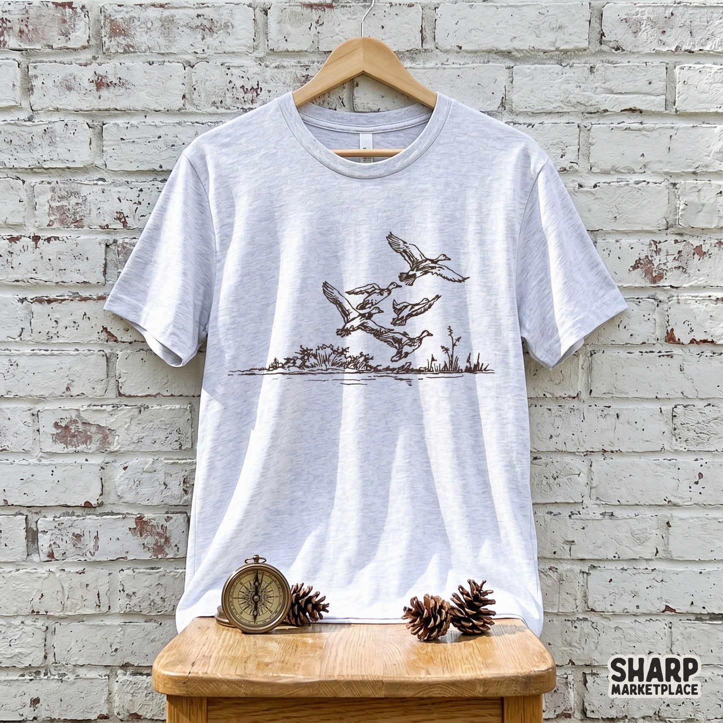 A light gray heather t-shirt featuring a detailed brown ink illustration of ducks in flight over a marsh, perfect for outdoor apparel and hunting gear.