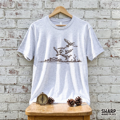 A light gray heather t-shirt featuring a detailed brown ink illustration of ducks in flight over a marsh, perfect for outdoor apparel and hunting gear.