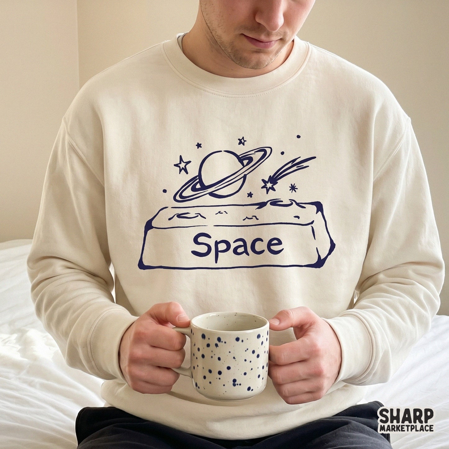Minimalist galaxy doodle design with a ringed planet and stars, shown as a dark blue graphic on a light-colored sweatshirt for apparel printing.