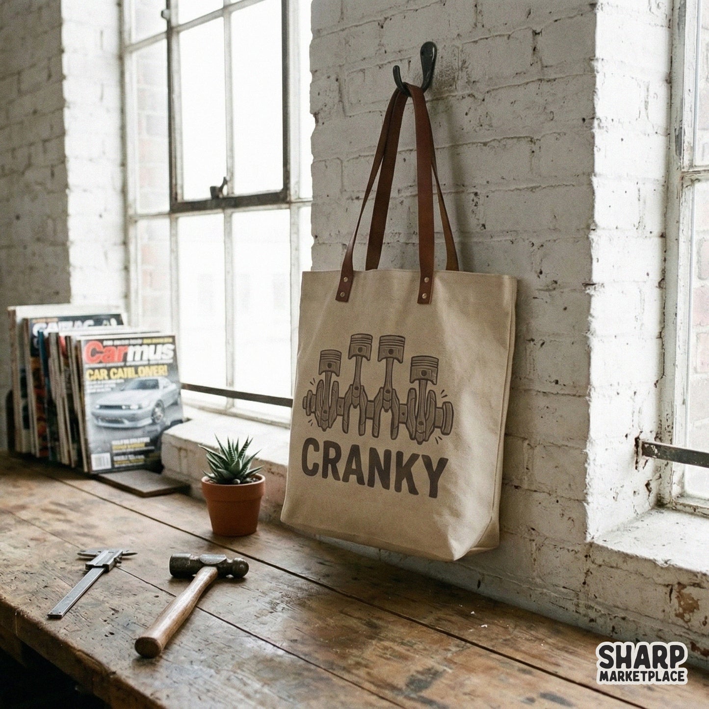 A canvas tote bag hanging on a wall, displaying a retro automotive design with four engine pistons and the text Cranky in a muted brown tone.