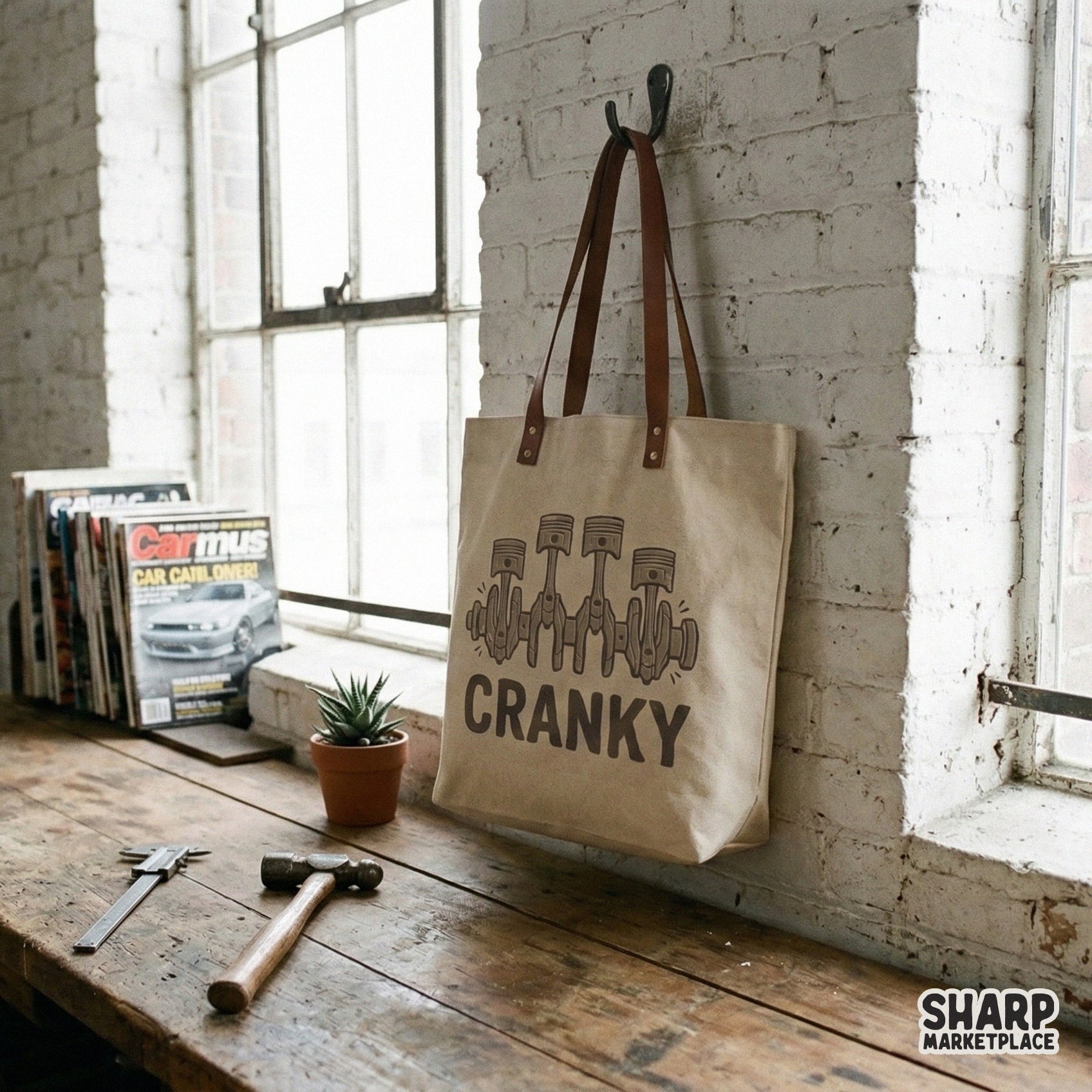 A canvas tote bag hanging on a wall, displaying a retro automotive design with four engine pistons and the text Cranky in a muted brown tone.
