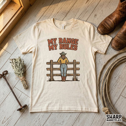 Sublimation design on a cream t-shirt featuring a cowgirl looking over a fence with the text My Range My Rules in a retro, distressed western font.
