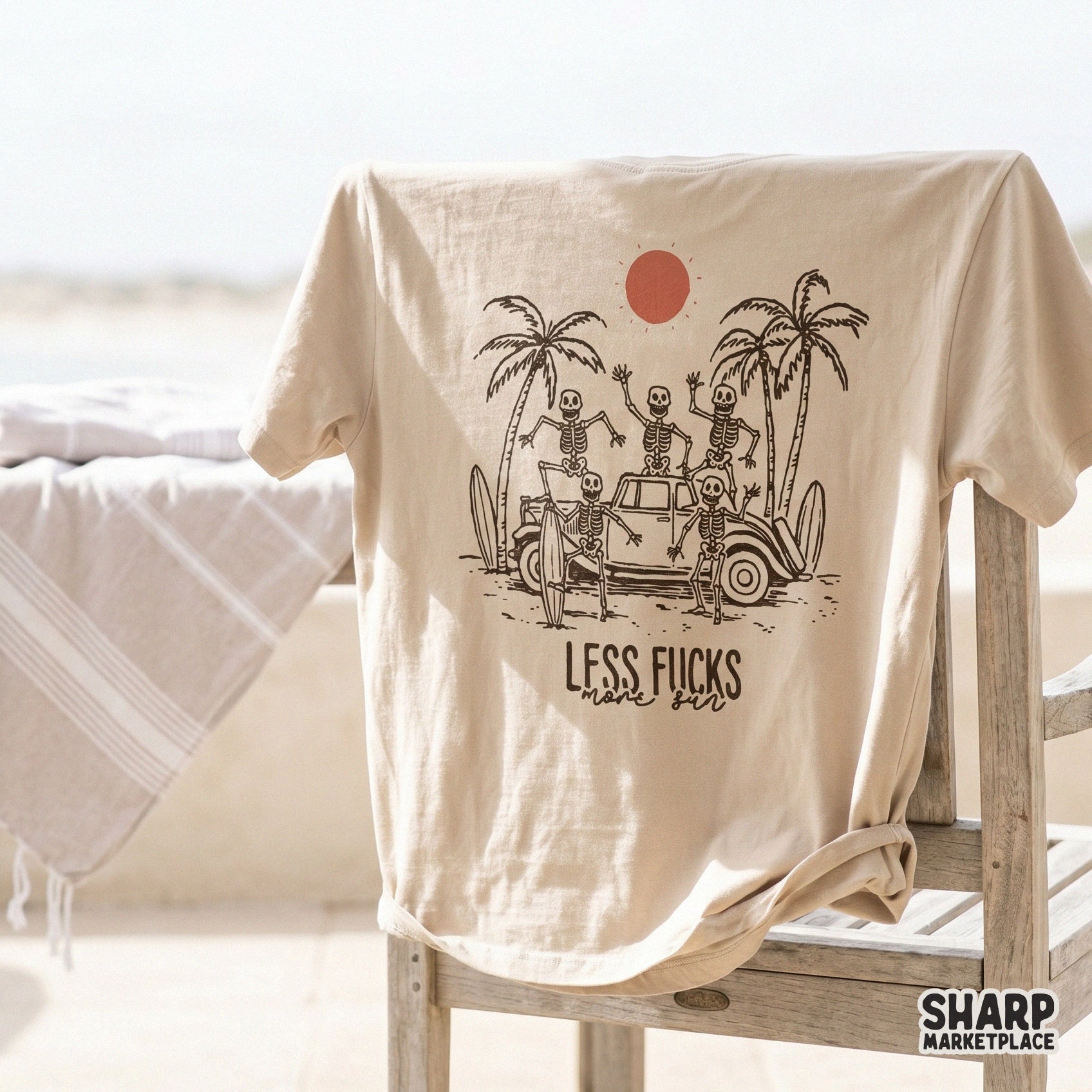A beige t-shirt mockup displaying a hand-drawn illustration of a skeleton surf crew at the beach with the quote Less Fucks More Sun in vintage lettering.