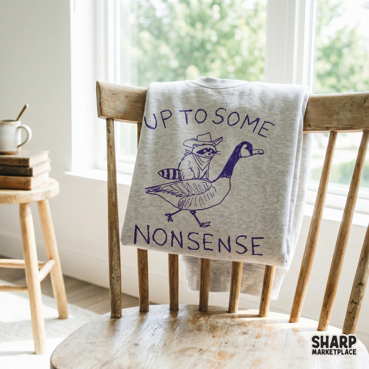 A purple ink style design featuring a raccoon in a cowboy hat riding a goose, displayed on a light gray sweatshirt for a trendy western animal aesthetic.