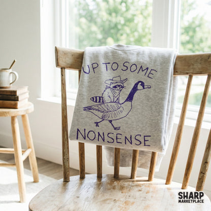 A purple ink style design featuring a raccoon in a cowboy hat riding a goose, displayed on a light gray sweatshirt for a trendy western animal aesthetic.