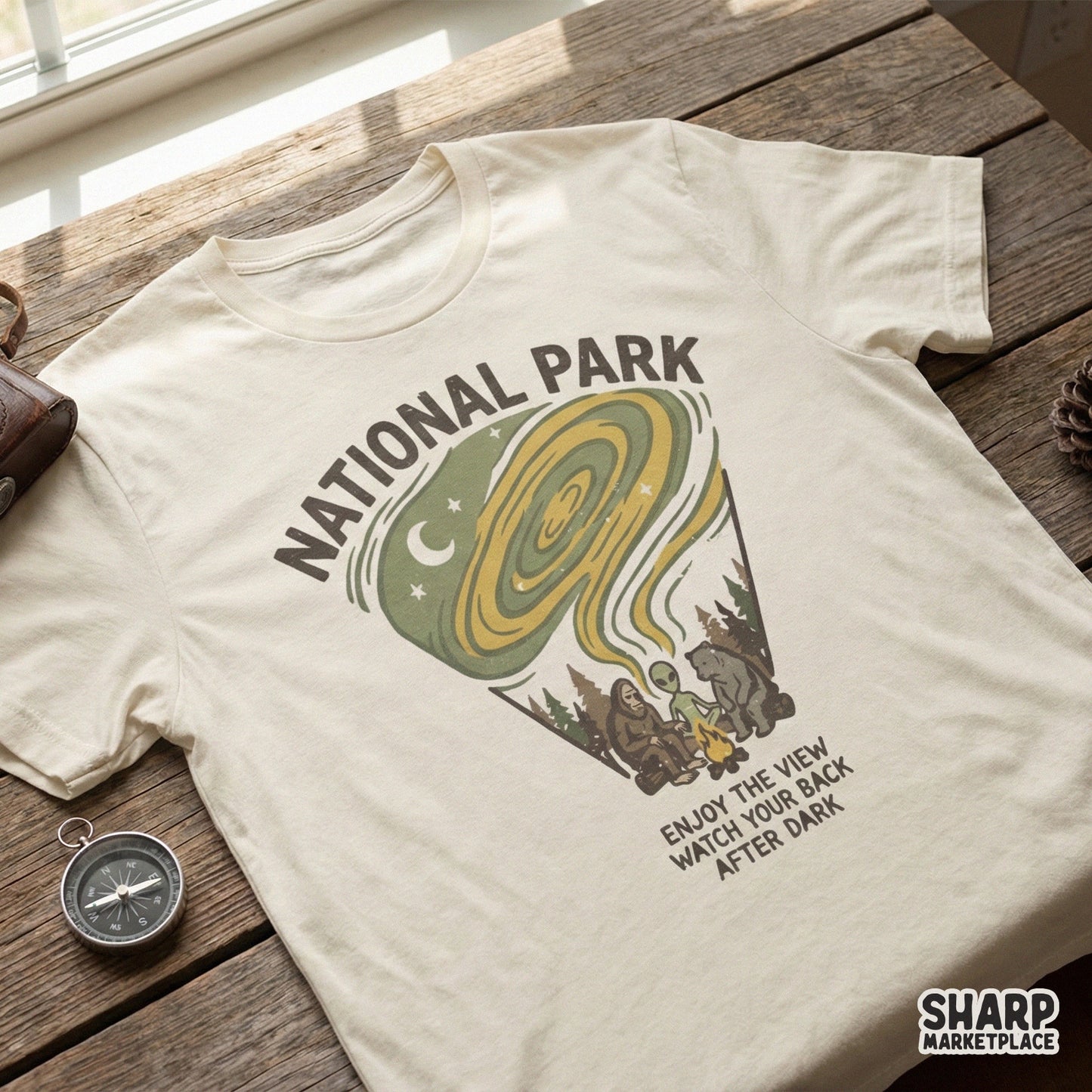 T-shirt mockup displaying a whimsical camping design with a forest creature, bear, and alien sitting at a campfire under a crescent moon and sage green sky.