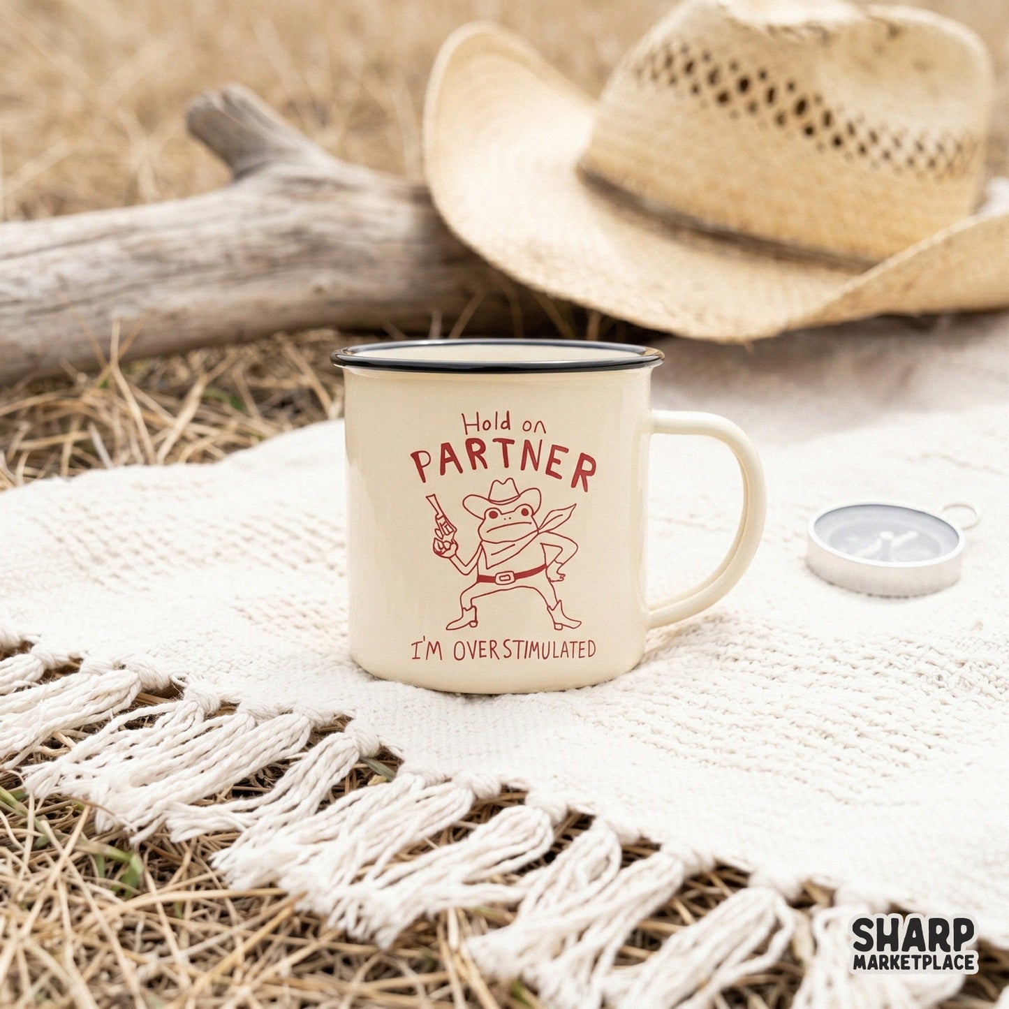 A cream-colored enamel mug featuring a red line art illustration of a cowboy frog with a gun and the quote Hold on Partner I’m Overstimulated.