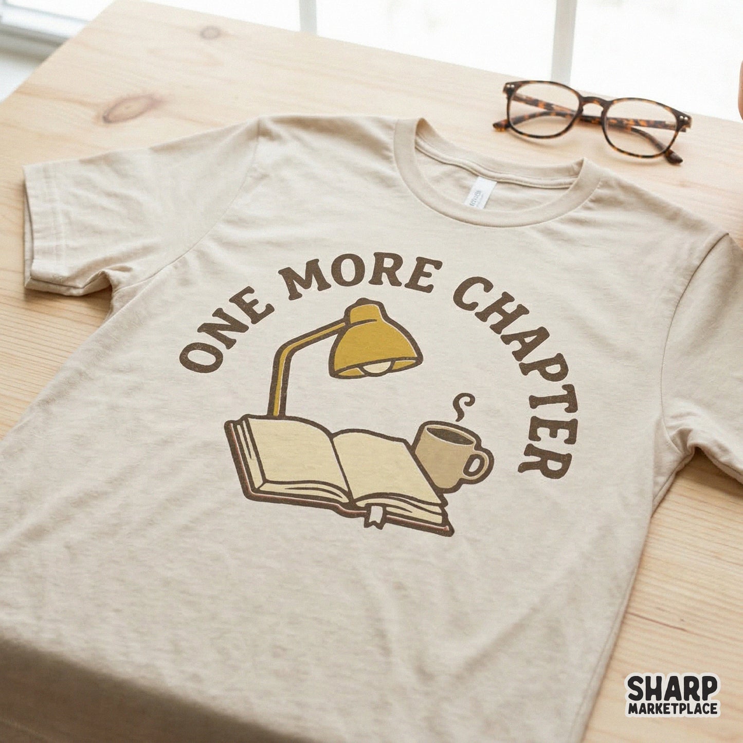 Sublimation design for apparel showing an open book, lamp, and coffee mug with One More Chapter text, displayed on a neutral colored t-shirt.