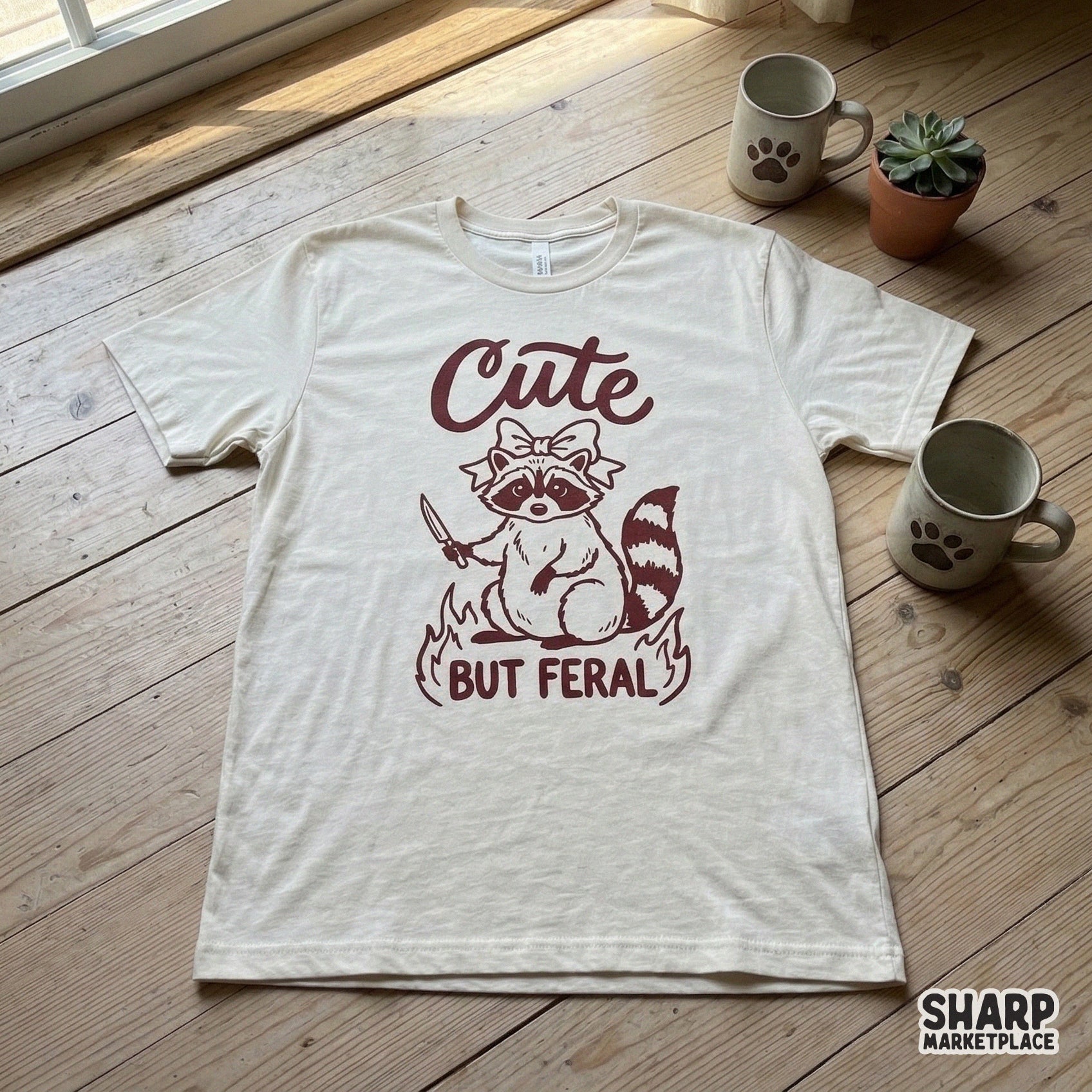 A cream-colored t-shirt featuring a maroon graphic of a mischievous raccoon with a bow and knife, framed by fire and the phrase Cute But Feral.