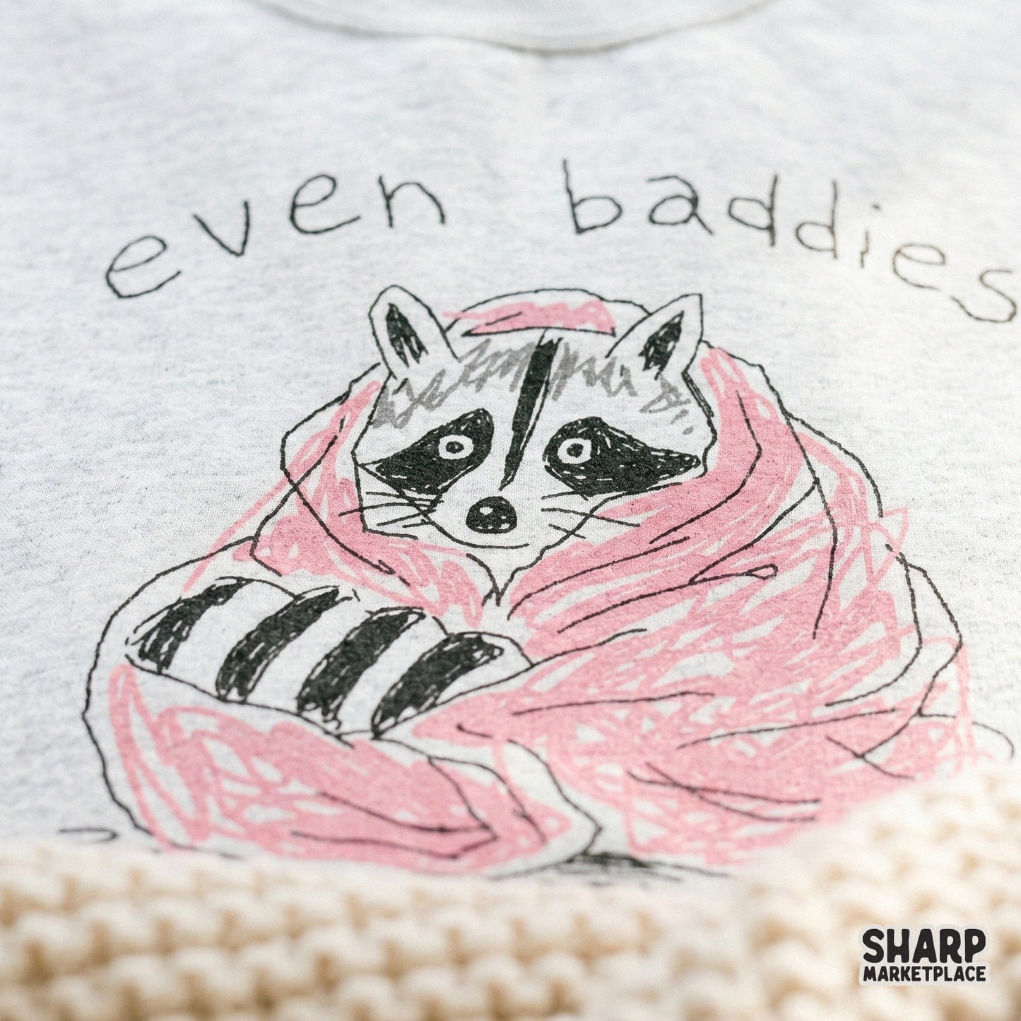 Funny raccoon graphic in a pink blanket with the quote Even Baddies Get Saddies, designed for print-on-demand shirts, tumblers, and vinyl decals.