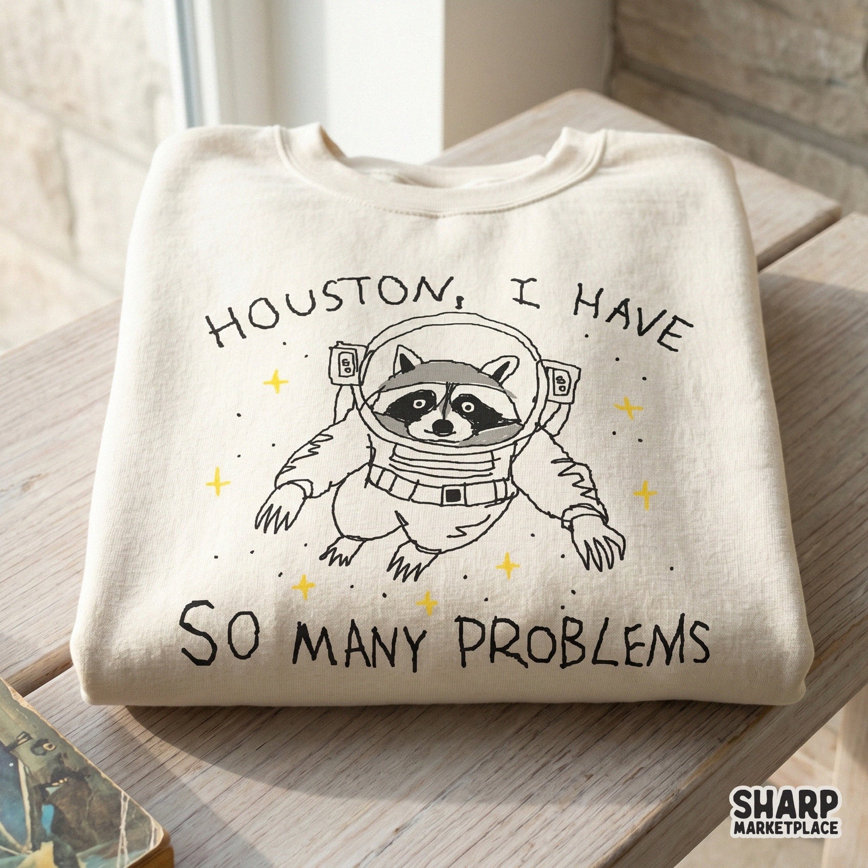 Digital design of a raccoon wearing an astronaut suit surrounded by stars with the quote Houston I Have So Many Problems for sarcastic apparel.