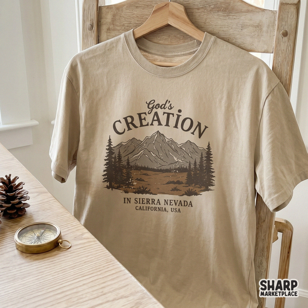 A beige t-shirt mockup displaying a rustic mountain landscape graphic with pine trees and faith-based text in earthy brown tones.
