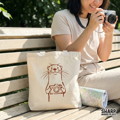 Hand-drawn otter mascot holding an SLR camera, featuring clean lines and a rustic aesthetic ideal for DIY print-on-demand shirts and laptop stickers.