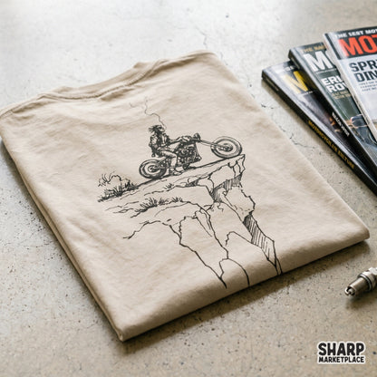 Vintage desert biker illustration featuring a rugged motorcycle rider on a canyon ledge, displayed as a black print on a tan t-shirt mockup.