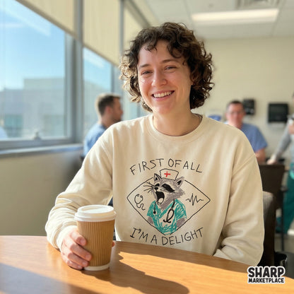 Digital sublimation design on a cream sweatshirt showing a chaotic raccoon nurse illustration with the witty quote First Of All I'm A Delight in black