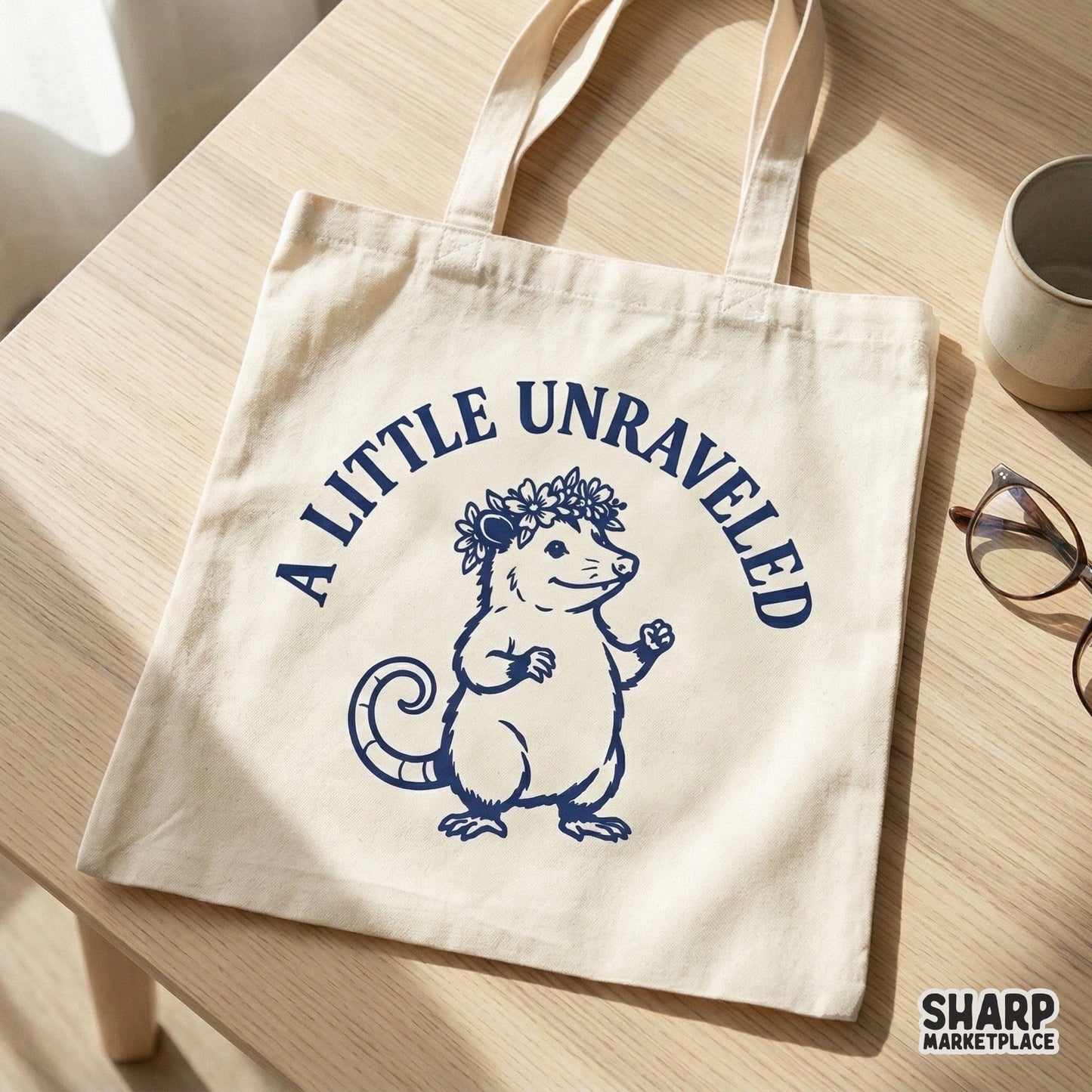 A canvas tote bag featuring a navy blue graphic of a whimsical opossum in a flower crown with the text A Little Unraveled for a cottagecore look.