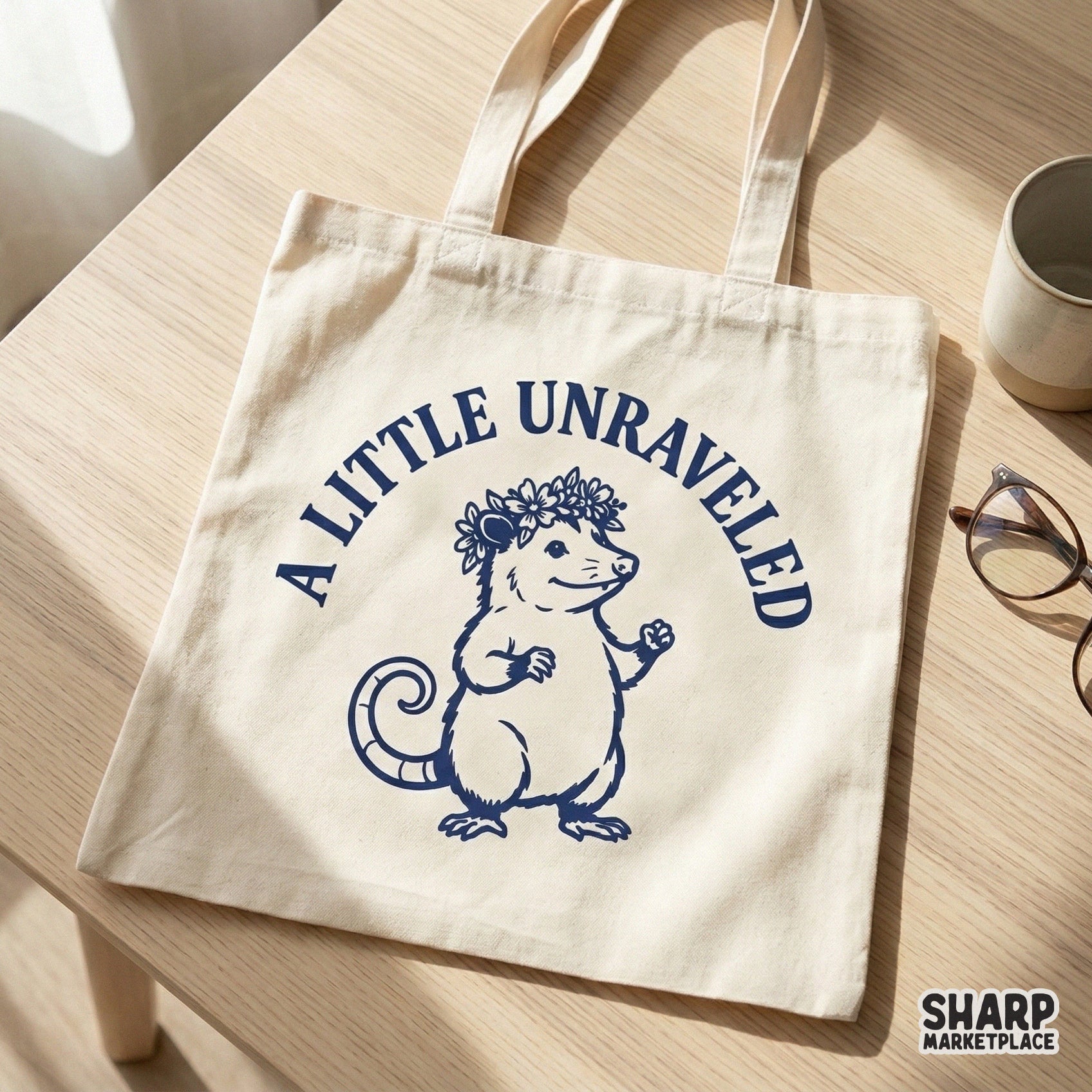A canvas tote bag featuring a navy blue graphic of a whimsical opossum in a flower crown with the text A Little Unraveled for a cottagecore look.