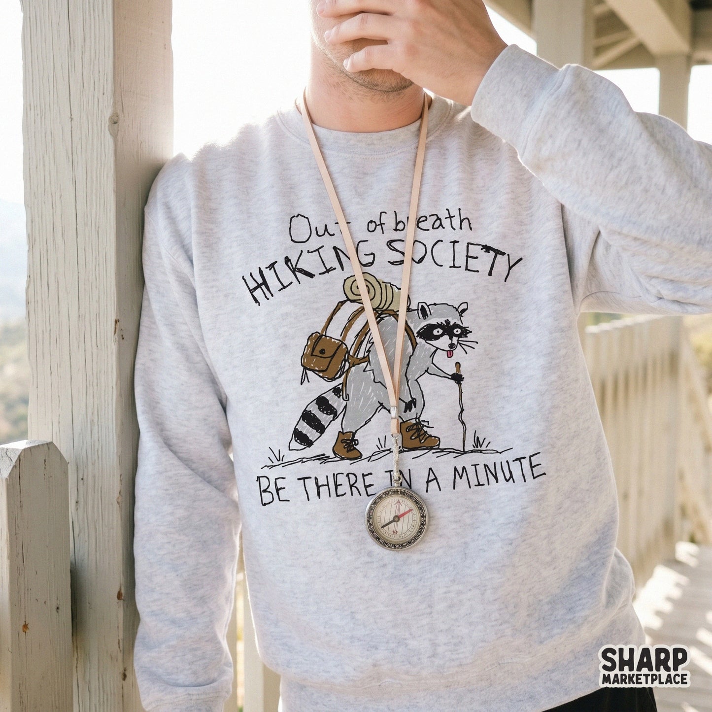 A light gray sweatshirt featuring a hand-drawn illustration of a tired raccoon hiker with a backpack and walking stick, captioned with humorous trail text.