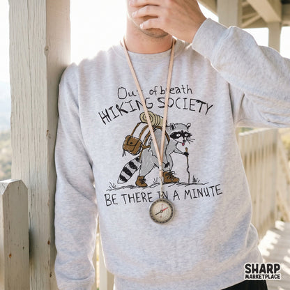 A light gray sweatshirt featuring a hand-drawn illustration of a tired raccoon hiker with a backpack and walking stick, captioned with humorous trail text.