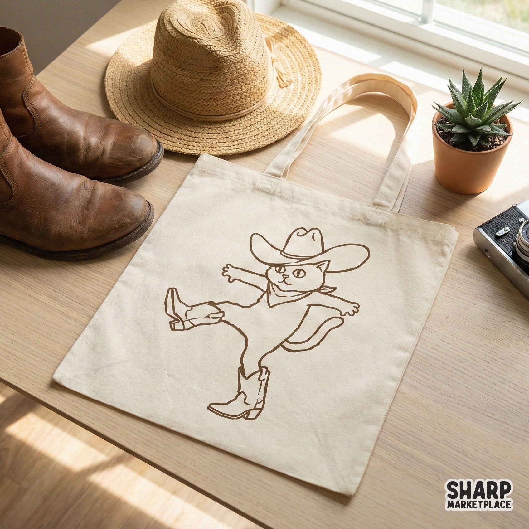 Brown line art illustration of a playful cowboy cat wearing western gear, displayed as a digital design on a canvas tote bag mockup.