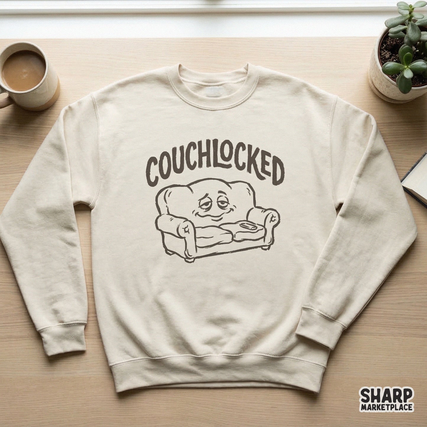 Beige sweatshirt mockup displaying a monochromatic black and white graphic of a sleepy, smiling sofa character with the word Couchlocked above it.