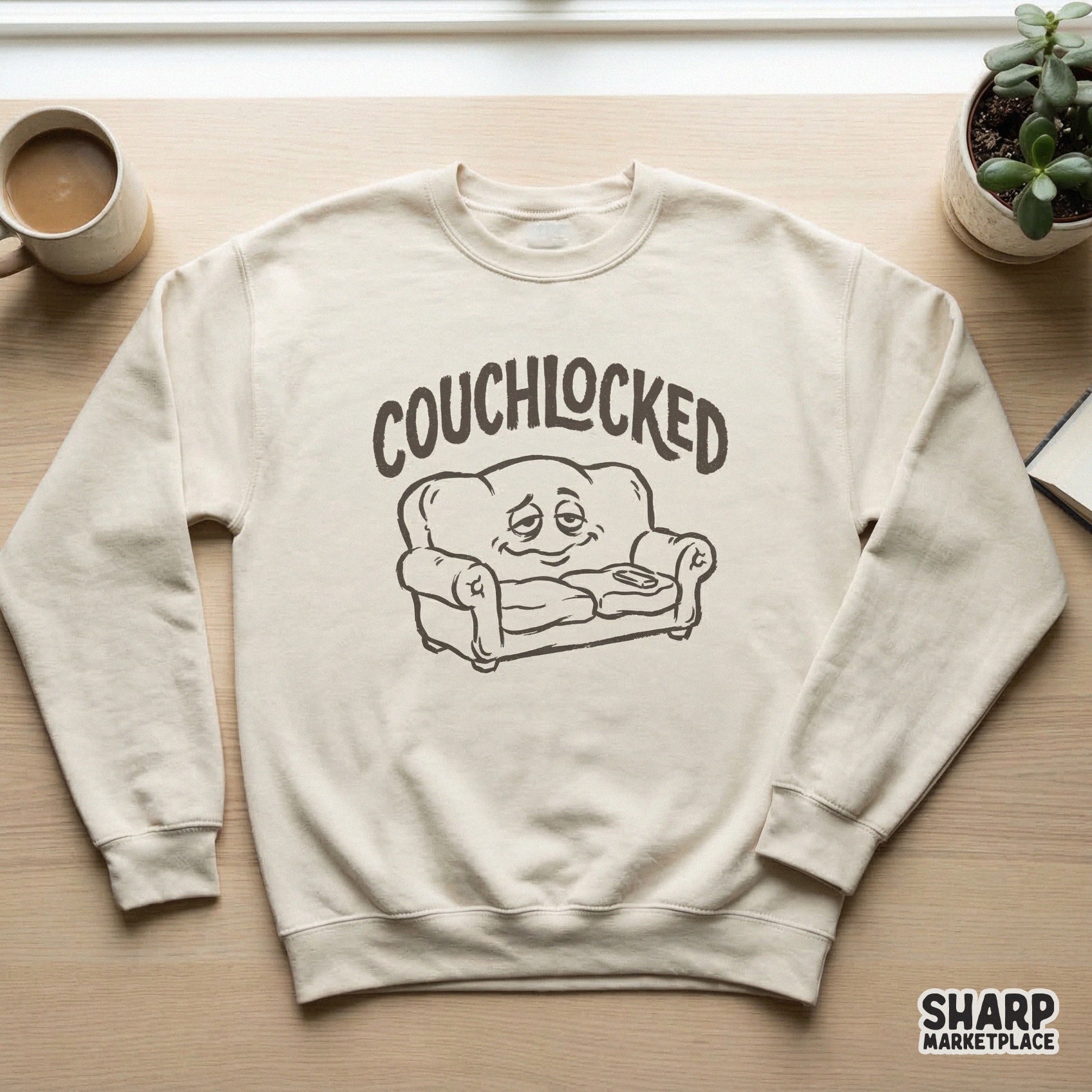 Beige sweatshirt mockup displaying a monochromatic black and white graphic of a sleepy, smiling sofa character with the word Couchlocked above it.