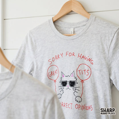 A light gray t-shirt featuring a hand-drawn white cat wearing sunglasses, holding red balloons, and displaying a sassy feminist quote in red sketch font.