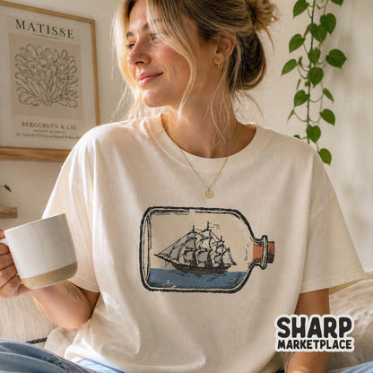 Retro-style nautical ship in a bottle graphic printed on a cream-colored t-shirt, showcasing a detailed sailing vessel on blue water.