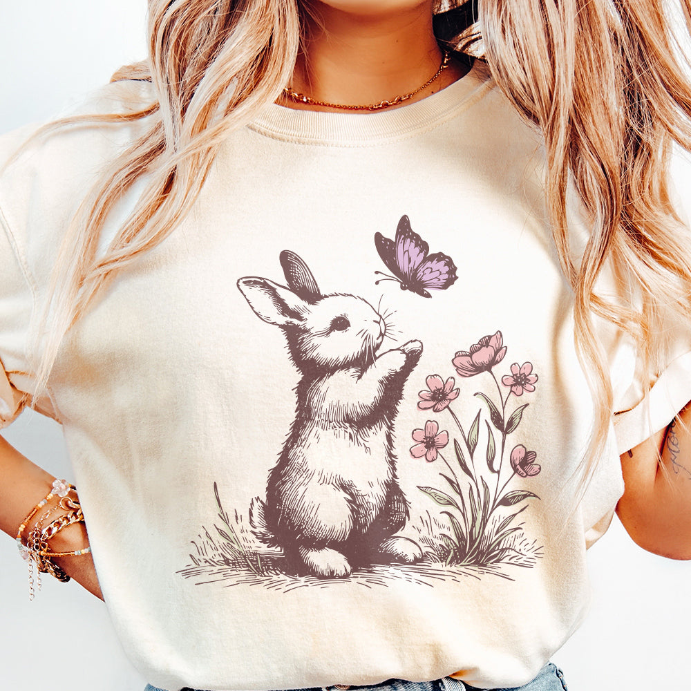 Cute rabbit reaching for a butterfly near pink flowers, shown as a vintage-style graphic on a cream-colored t-shirt for apparel printing.
